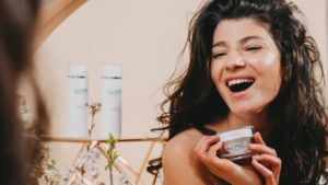A woman laughs while holding KONU cosmetics in front of a mirror.