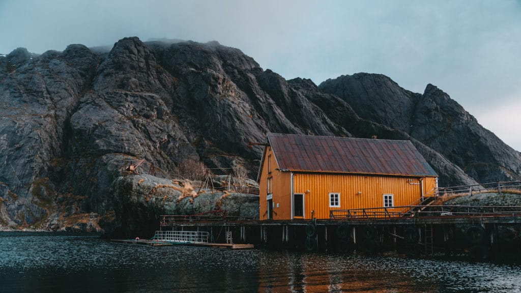 Orangenes Holzhaus am Wasser im Nusfjord mit Bergen im Hintergrund