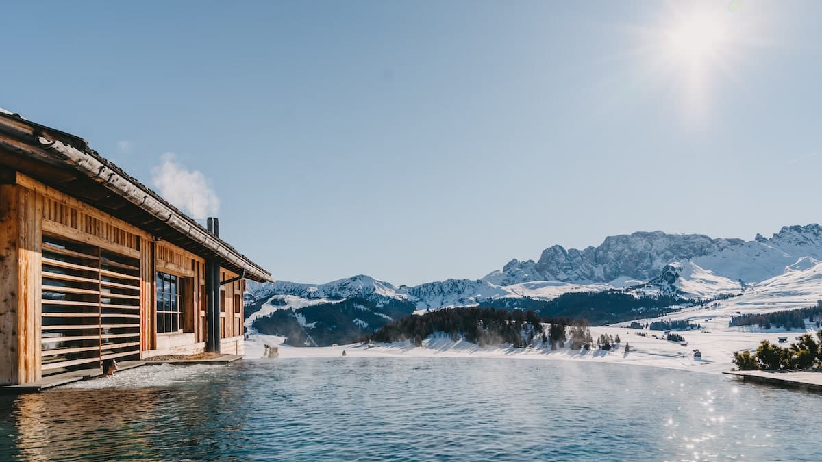 A wooden cabin with a hot tub in front of snowy mountains.