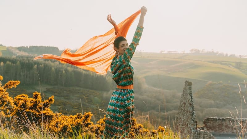 A woman in an orange dress looks adorable as she stands on a hill.